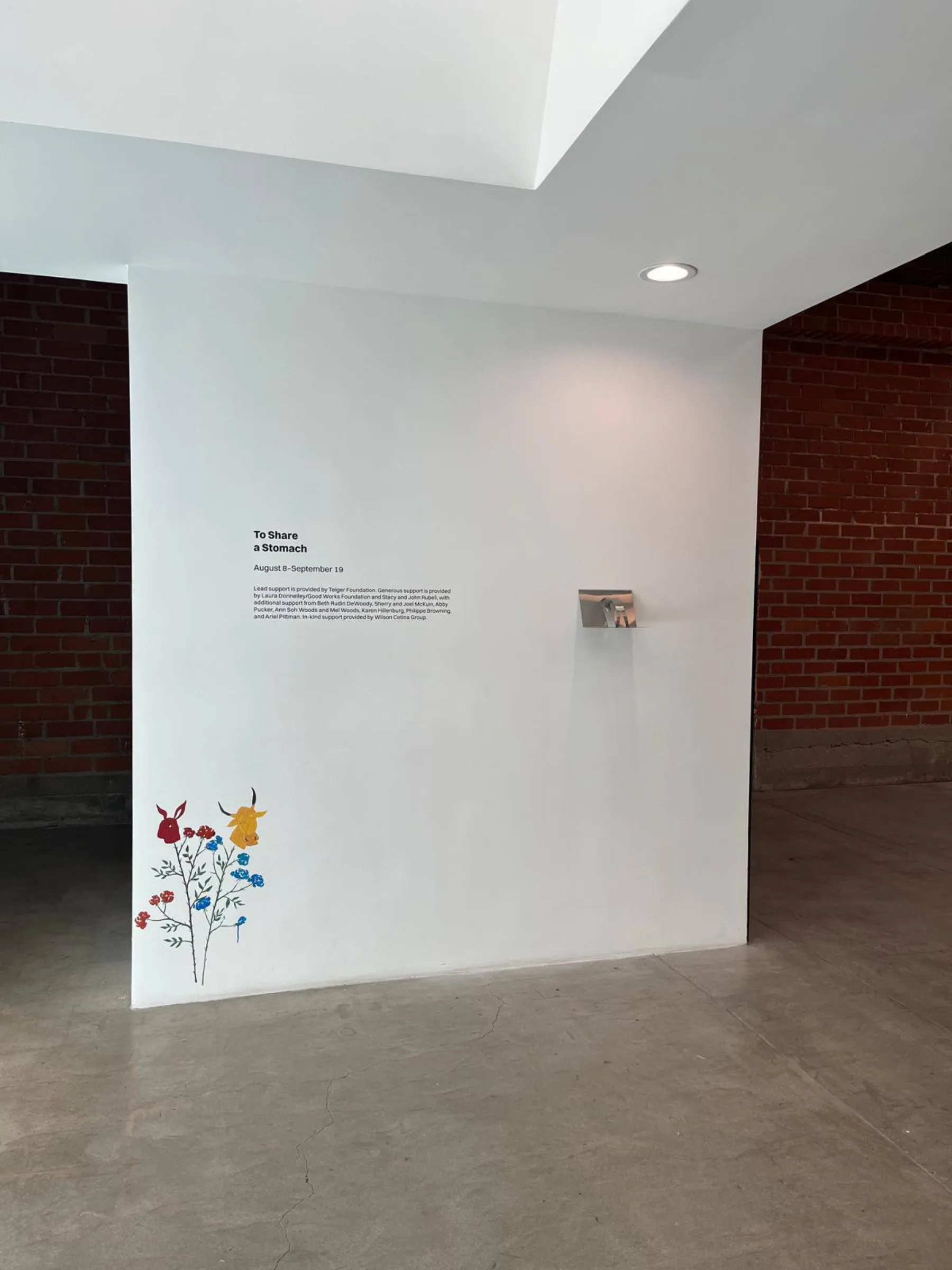 Wall text for To Share a Stomach beside a small painted mural detail.