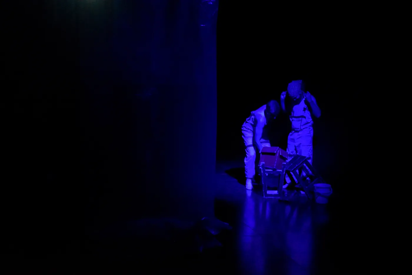 Two performers in blue light, bent over a small red house on a stool.