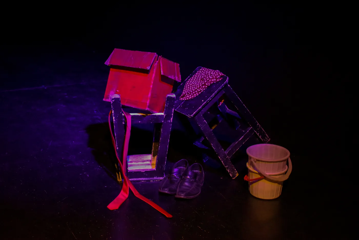 A small red house, a pair of shoes, and a bucket arranged on a dark stage under coloured light.