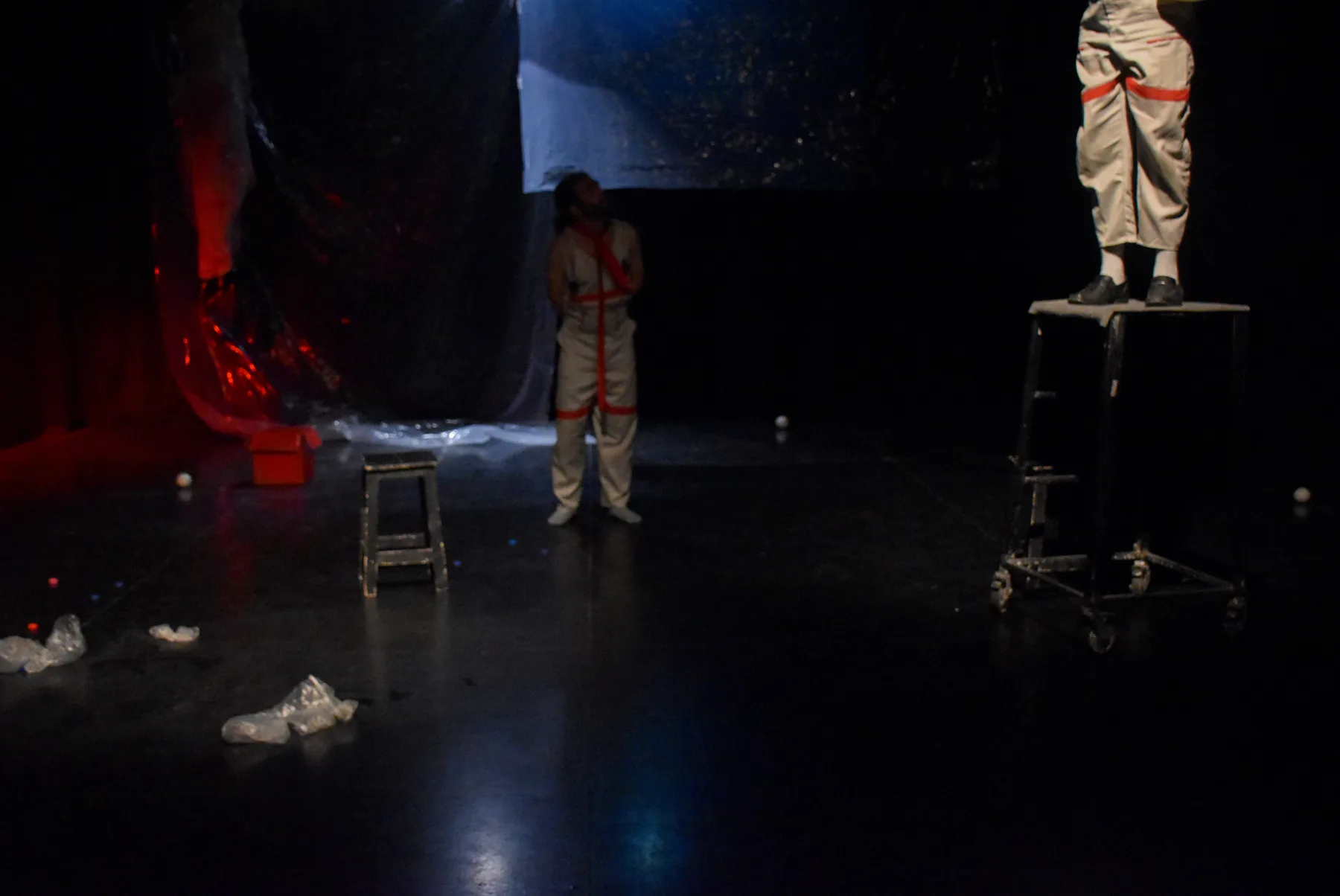 A performer stands beneath a projected image while another stands on a raised platform, red fabric spilling across the dark stage.